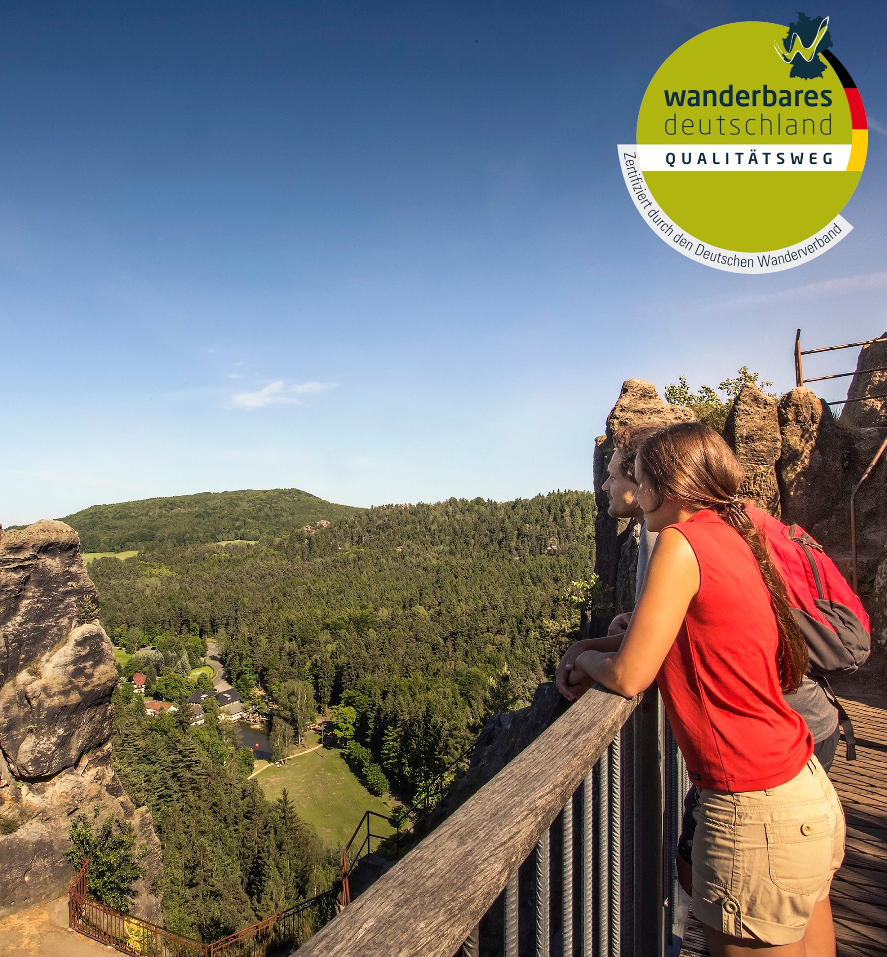 Zwei Personen stehen auf einer Aussichtsplattform und schauen auf den Kurort Jonsdorf. In der Ecke befindet sich das Logo Qualitätsweg Deutschland.